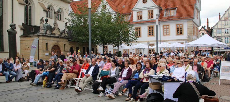 Menschen sitzen auf Stühlen auf einem Platz und schauen sich ein Open Air Konzert an. 