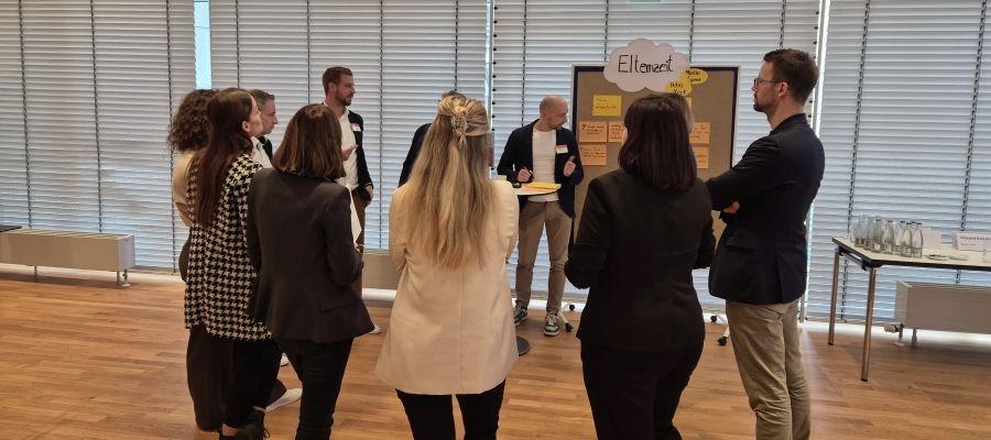 Ein Personenkreis steht um ein Flipchart herum und ist im Austausch zu dem Thema Elternzeit. Quelle: Linda Lüdeke