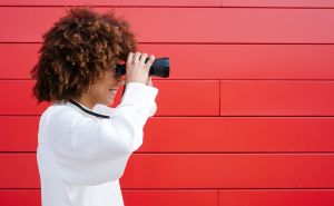 Frau schaut durch ein Fernglas vor roter Wand. - Quelle: DSV