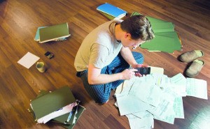 Ein Student sitzt umgeben von Dokumenten auf dem Boden und tippt auf einem Taschenrechner.