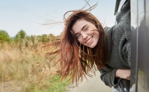 Frau mit braunen Haaren hält ihren Kopf aus dem fahrenden Auto und lacht in die Kamera